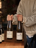 A person in a beige and black striped sweater holds a bottle of wine next to two other Pooley wine bottles on a wooden table in a cozy indoor setting.