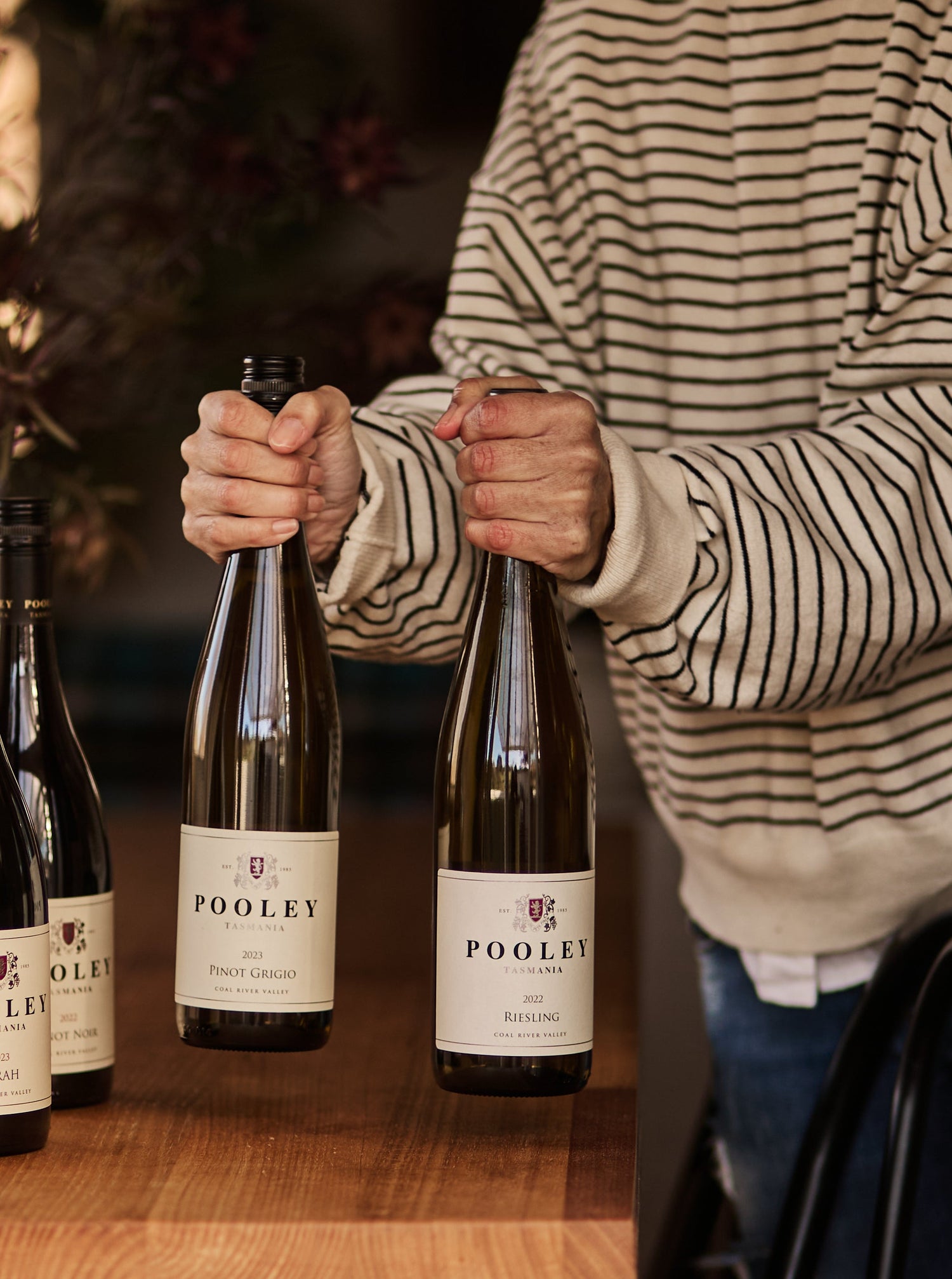 A person in a beige and black striped sweater holds a bottle of wine next to two other Pooley wine bottles on a wooden table in a cozy indoor setting.
