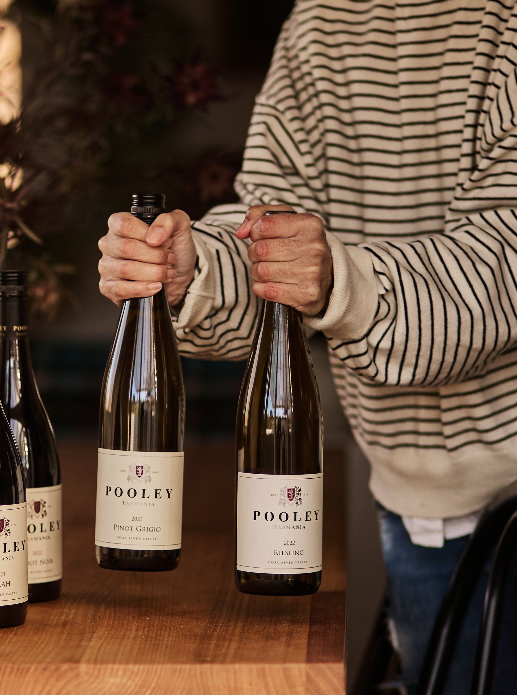 A person in a beige and black striped sweater holds a bottle of wine next to two other Pooley wine bottles on a wooden table in a cozy indoor setting.